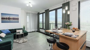 A modern living room with a green sofa, a dining area with a wooden table, and large windows offering a city view. - Warsaw Flat With Balcony (Warsaw)