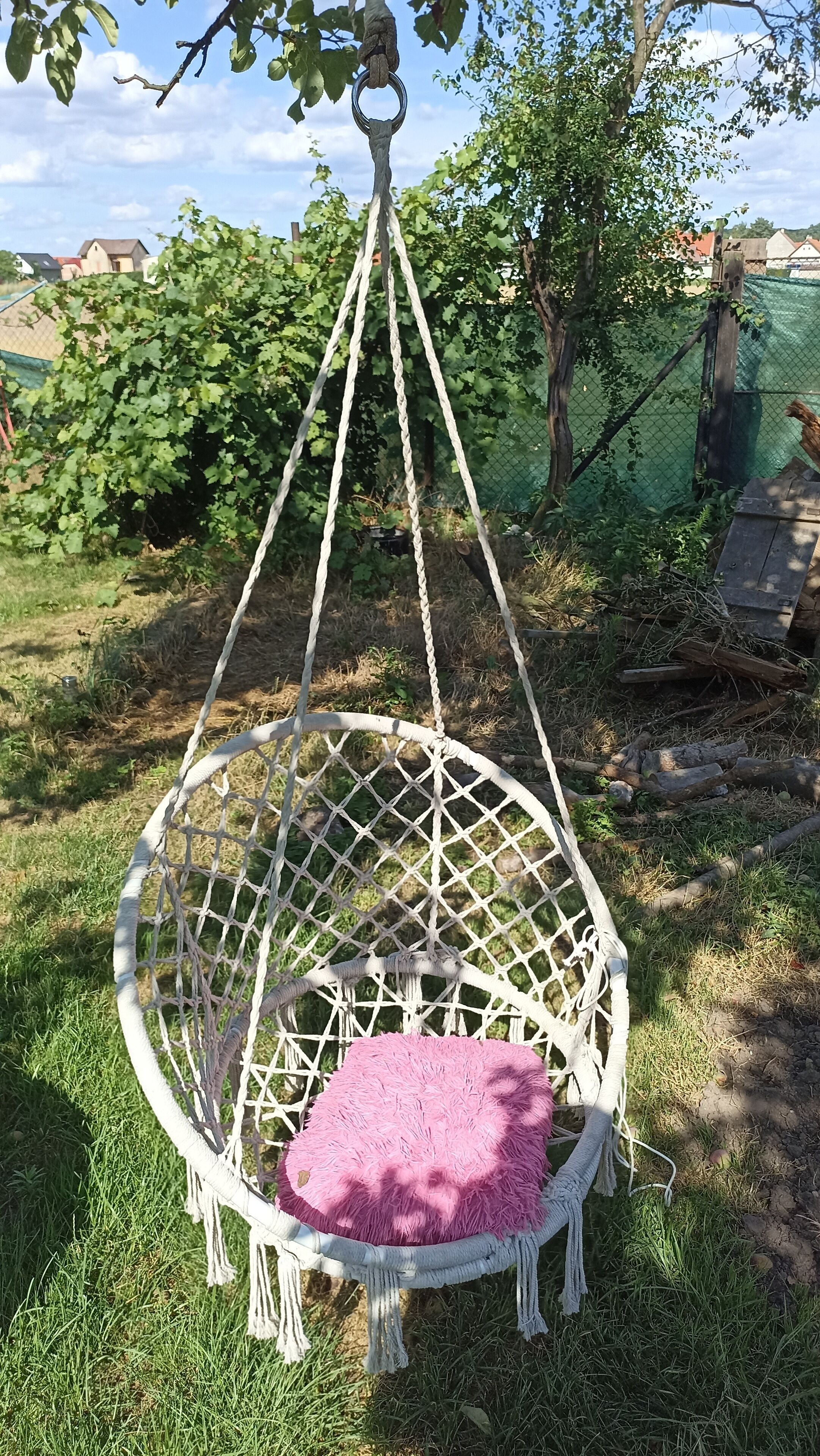 Ein weißer Hängestuhl mit rosa Kissen in einer grünen Außenanlage.
