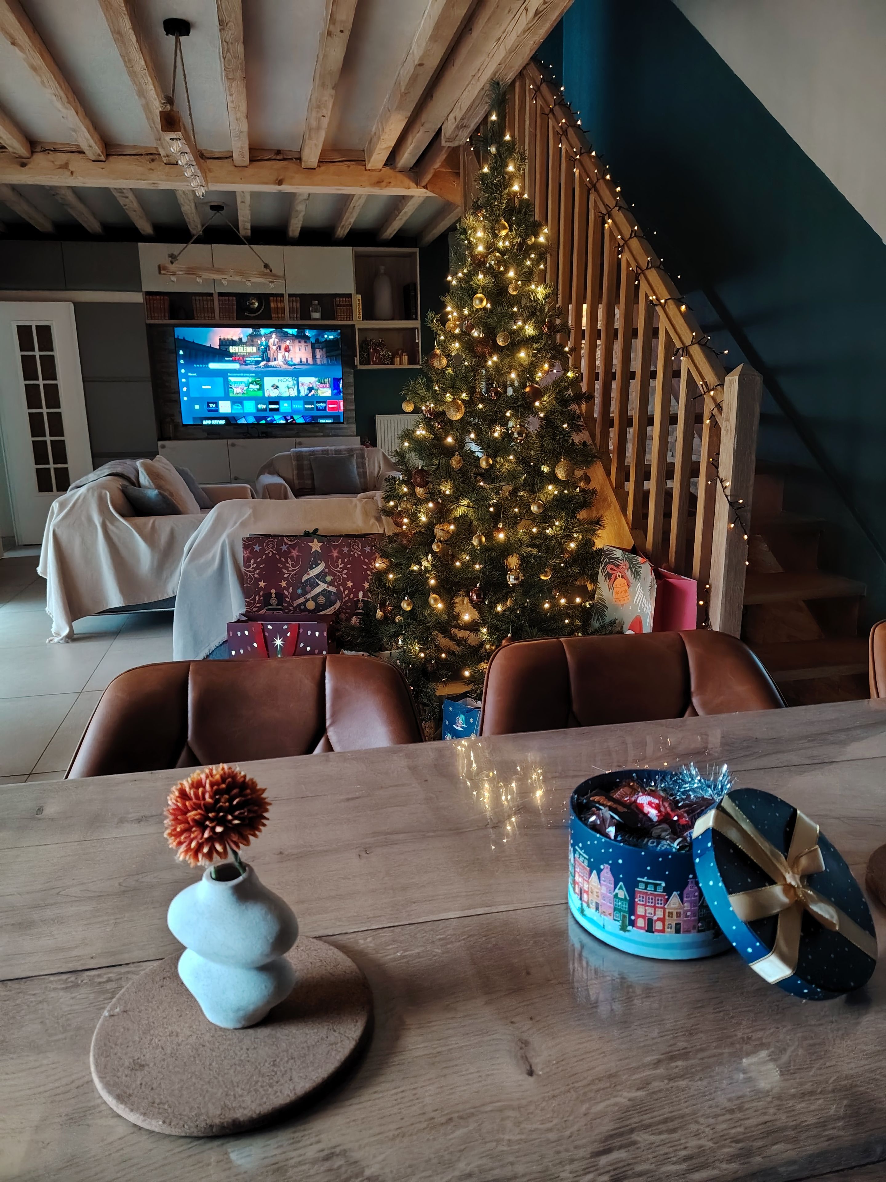 Un salon chaleureux avec un sapin de Noël décoré, une télévision et une table à manger sur laquelle sont disposés des cadeaux.