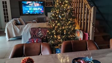 Un salon chaleureux avec un sapin de Noël décoré, une télévision et une table à manger sur laquelle sont disposés des cadeaux.
