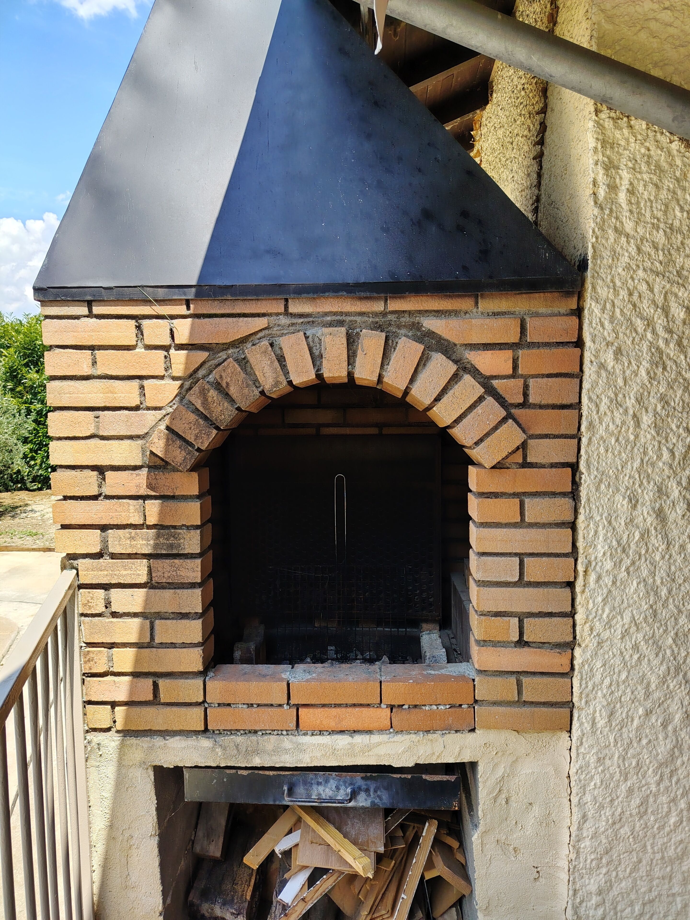 Un four extérieur en briques, doté d’un toit noir, d’un foyer et d’une pile de bois de chauffage en dessous.