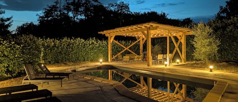 A wooden gazebo with seating, a swimming pool, and a paved deck area.