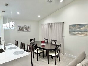 A dining area with a round table, six chairs, a white countertop with a sink, pendant lights, a painting of a landscape, and a window with curtains. - Cozy 2-bedroom pet friendly family home in central Margate with pool access. (Margate)