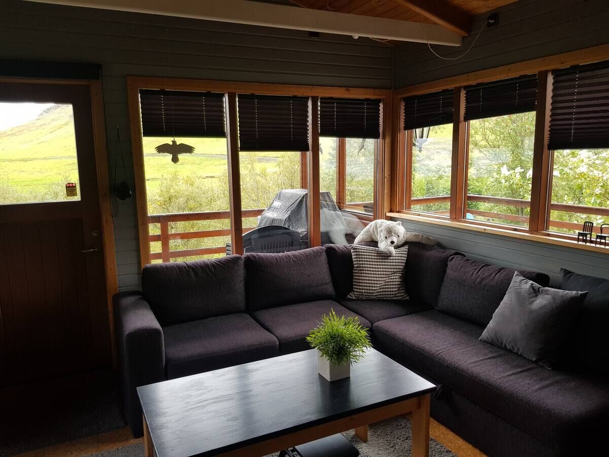 A cozy room with a sectional sofa, a small table with a plant, and large windows with blinds.