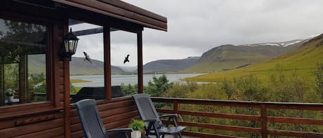 Um deck de madeira com duas cadeiras e uma mesa, com vista para um lago e colinas.
