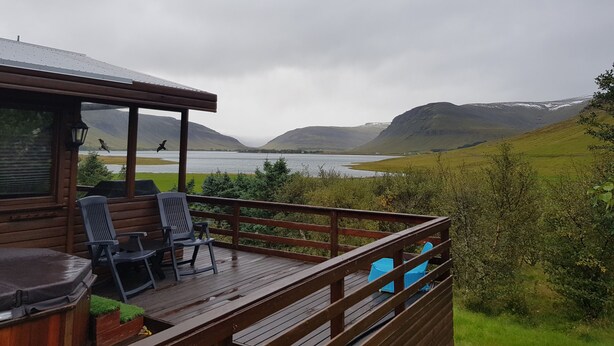 Una terraza de madera con mobiliario exterior, con vistas a un lago y montañas.