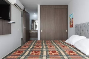 Ein Hotelzimmer mit einem gemusterten Teppich, einem Bett mit weißer Bettwäsche, einem grau gepolsterten Kopfteil und einer Holztür.