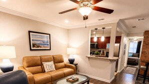 Un salon avec un canapé en cuir marron, un fauteuil en cuir bleu, une table basse en verre et un ventilateur de plafond.