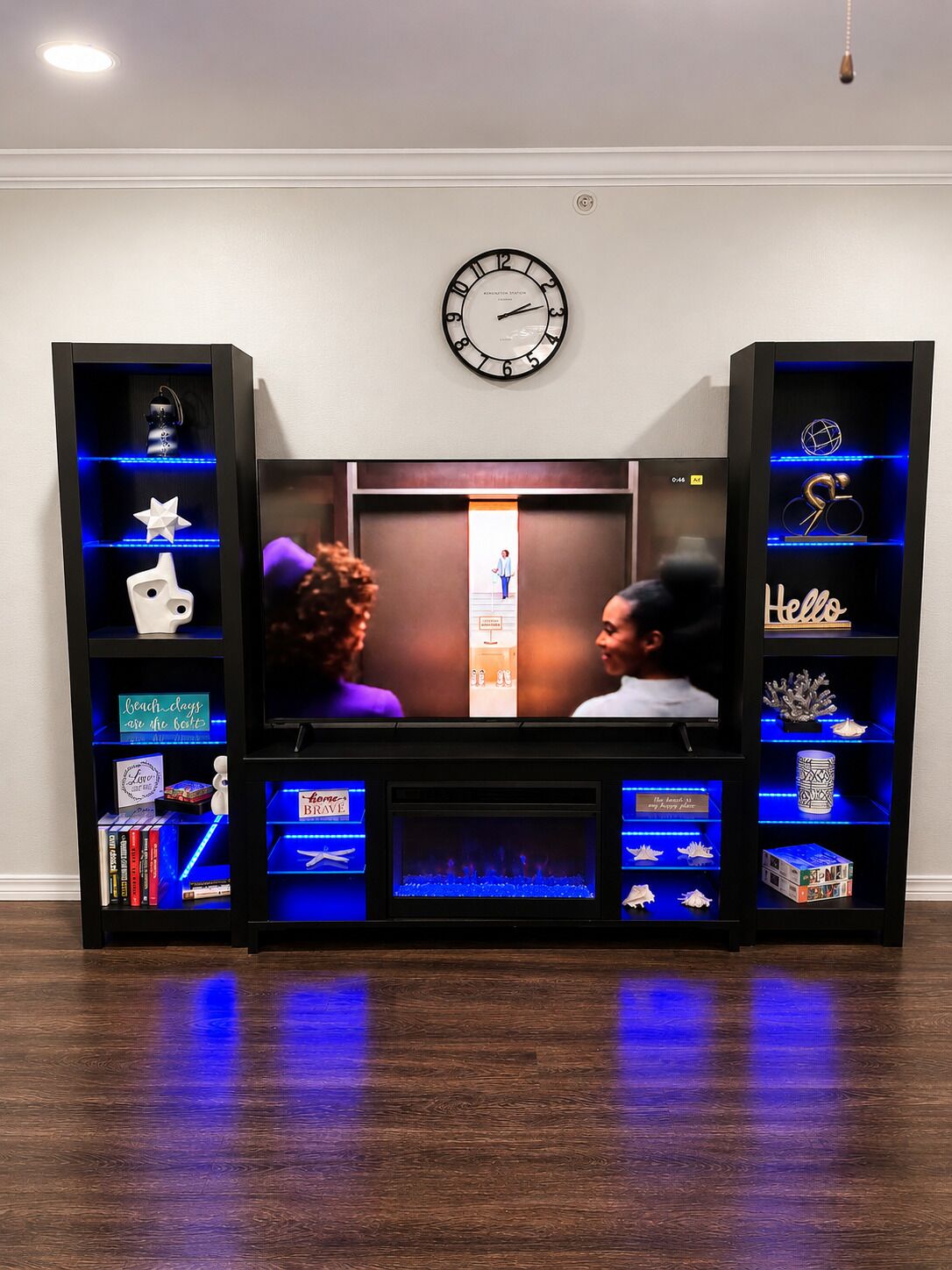 A living room with a television, two black shelving units with decorative items, a wall clock, and a wooden floor.