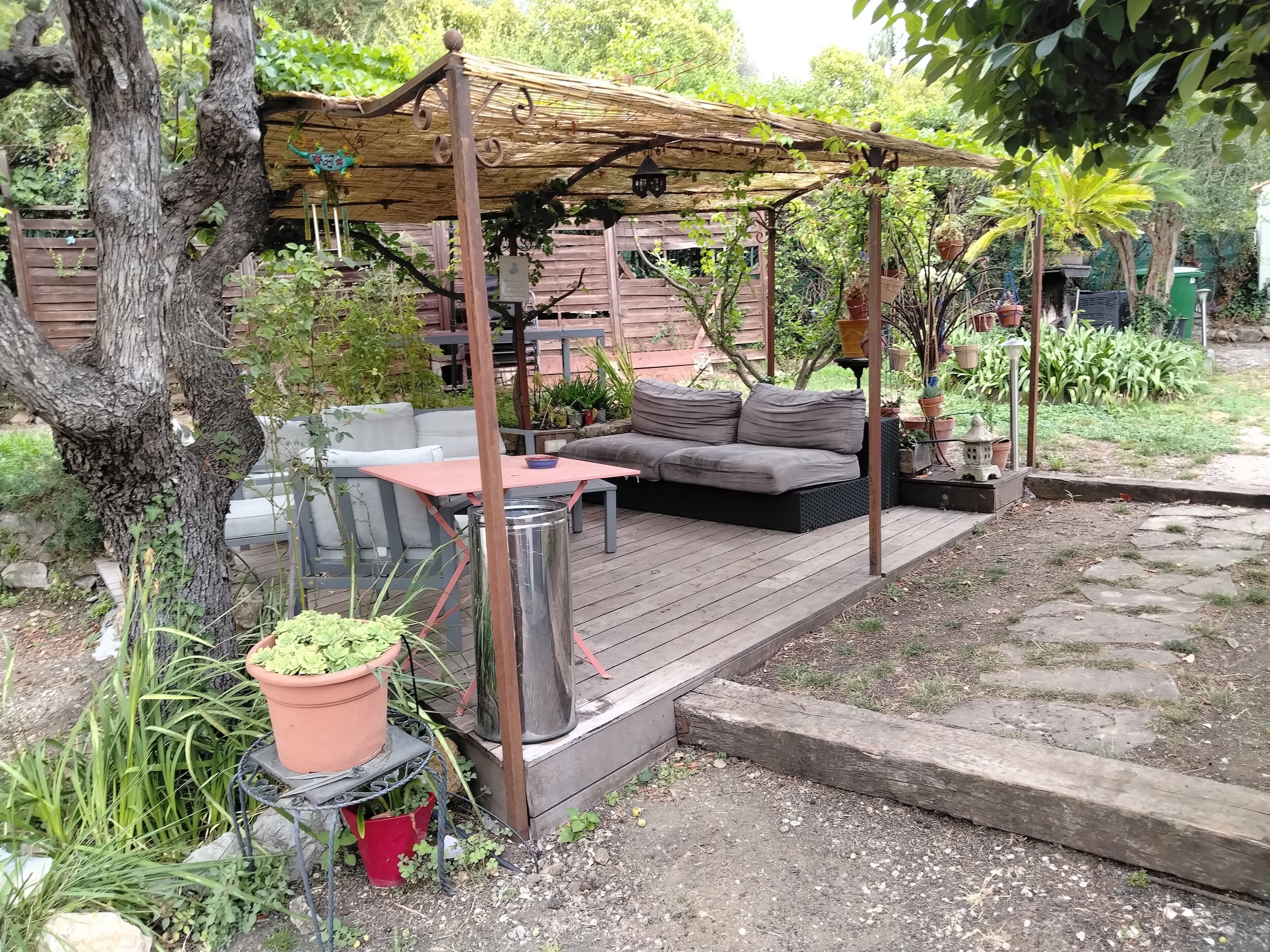 Ein überdachter Außenbereich mit Sitzgelegenheiten, inklusive Tisch und Sofa, umgeben von Topfpflanzen und einer Holzterrasse.