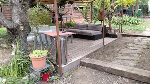 Un espacio de estar techado al aire libre con una mesa y un sofá, rodeado de plantas en macetas y una terraza de madera.