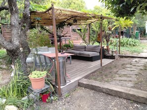 Ein überdachter Außenbereich mit Sitzgelegenheiten, inklusive Tisch und Sofa, umgeben von Topfpflanzen und einer Holzterrasse.