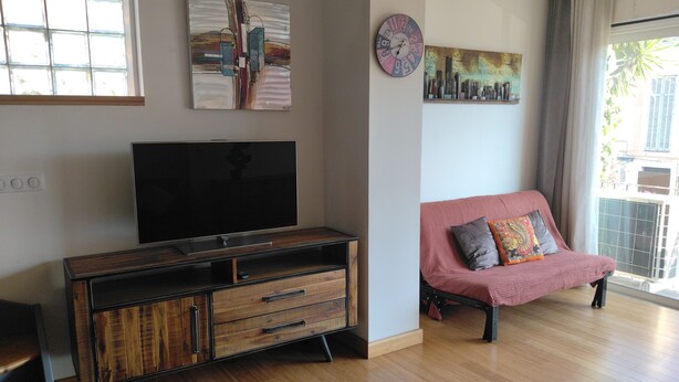 Una sala de estar con una televisión de pantalla plana sobre un mueble de madera, un sofá rojo con cojines y un cuadro con una vista urbana en la pared.