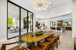 A modern interior with a dining area featuring a glass-top table and wooden chairs, a kitchen with a large island, and a living area with a sofa and a coffee table. - Brand-New, Spacious Desert Oasis - Pool, Spa & Mountain Views (La Quinta)