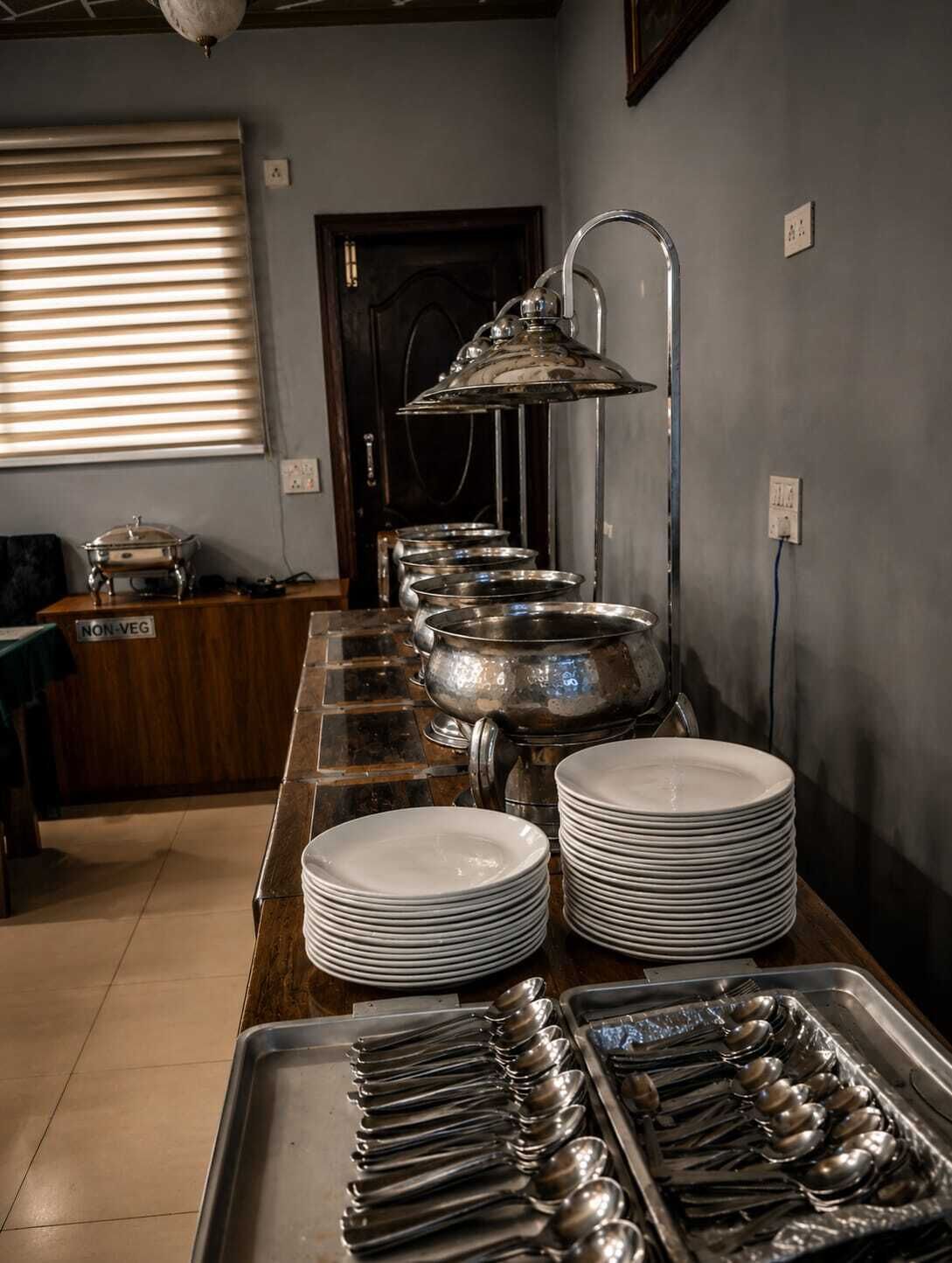 A buffet setup with stacked plates and serving utensils, a non-vegetarian sign, and a window with blinds.