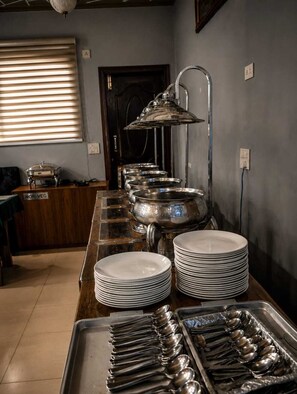 Buffet montado com pratos empilhados e utensílios de servir, placa indicando que não é vegetariano, e uma janela com persianas.