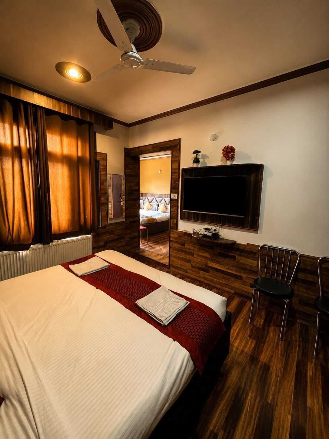 Habitación de hotel con una cama, un televisor de pantalla plana en la pared, un ventilador de techo y suelo de madera.