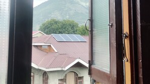 Ventana con vista a una casa de techo rojo y paneles solares, una montaña al fondo y un cielo parcialmente nublado.
