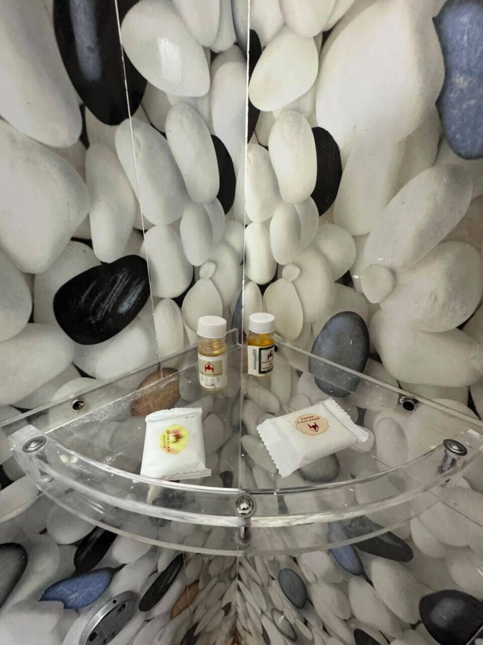 Una mesa redonda de vidrio con una toalla blanca y dos botellas, rodeada por un muro de guijarros blancos y negros.