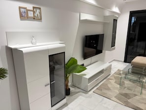 Una sala de estar moderna con un televisor de pantalla plana montado en la pared, un mueble blanco para entretenimiento y un aparador de vidrio con una planta.