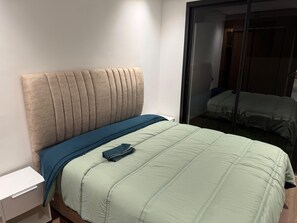 Una cama bien hecha con cabecera, mesita de noche y un espejo que refleja una habitación con cama.