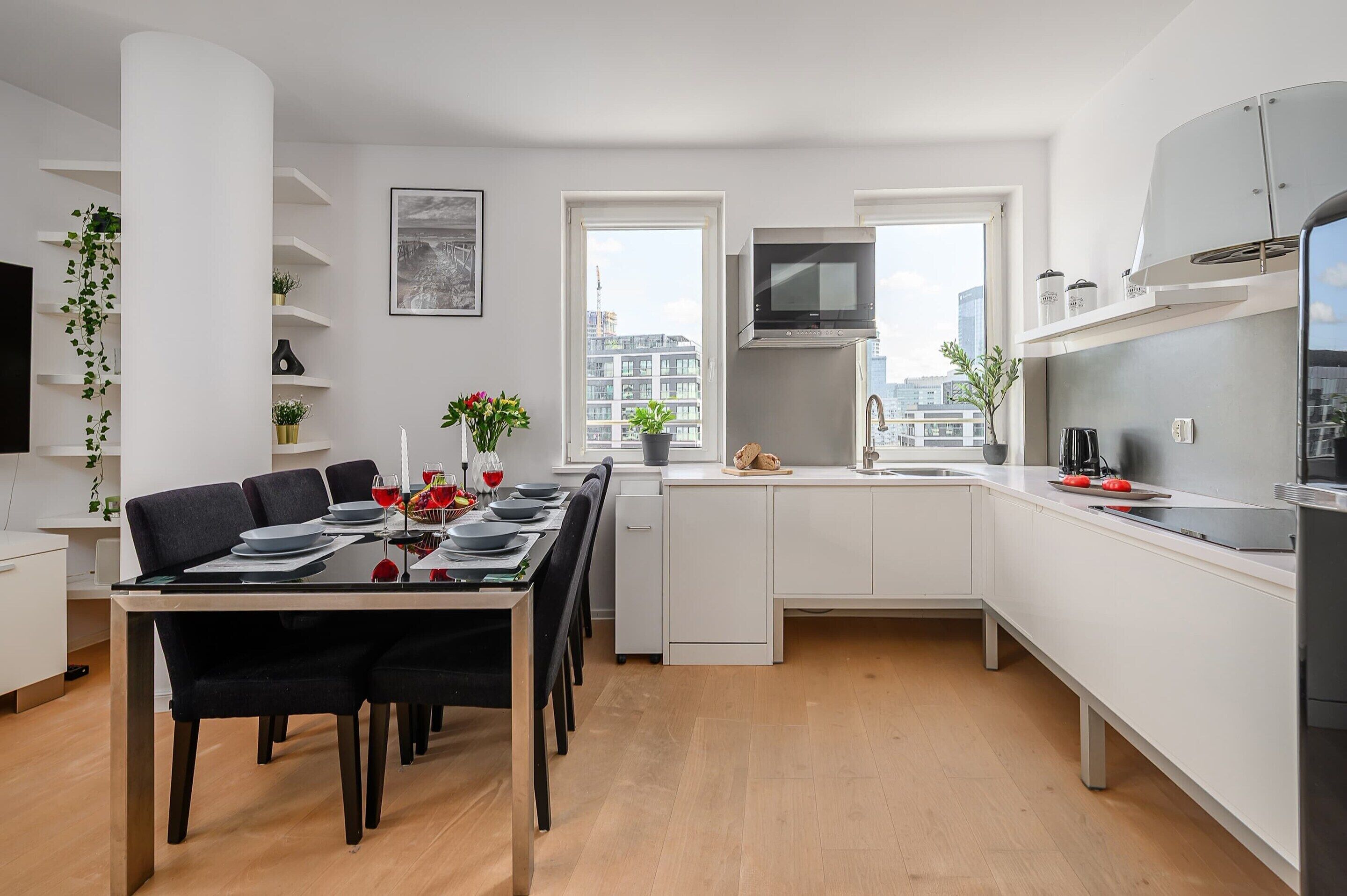 A modern kitchen with white cabinetry, a dining area with a table set for four, and a view of the city through large windows.