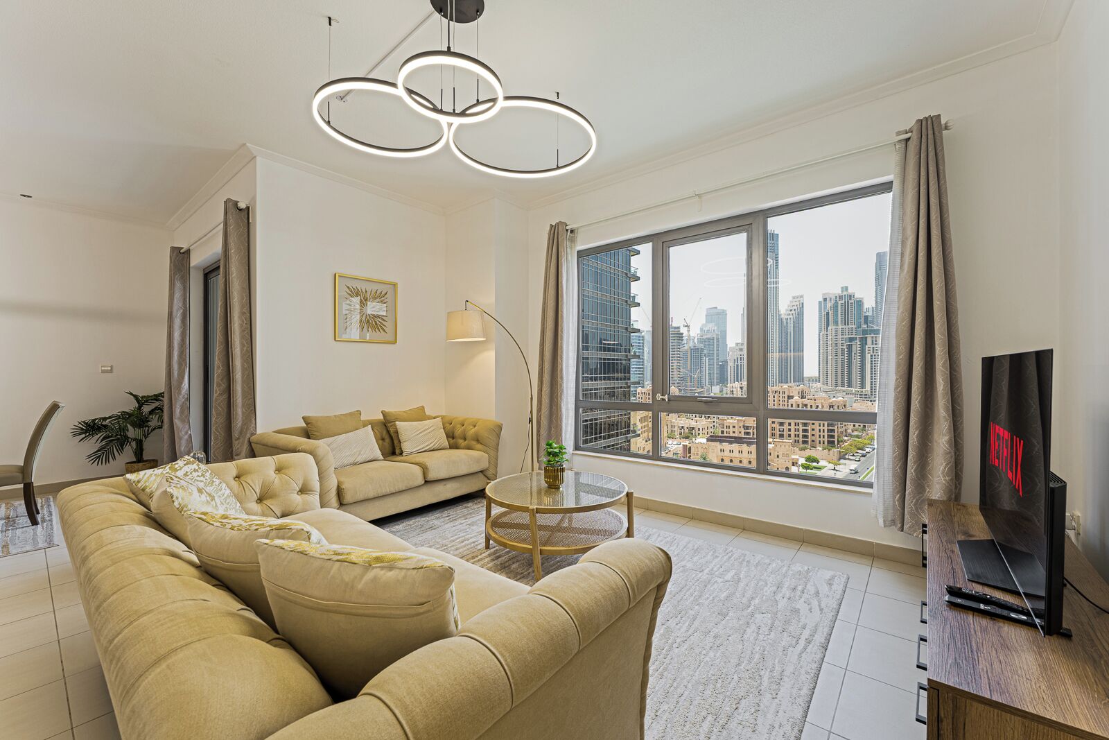 A modern living room with a sofa, a round coffee table, a TV, and a large window with a city view.