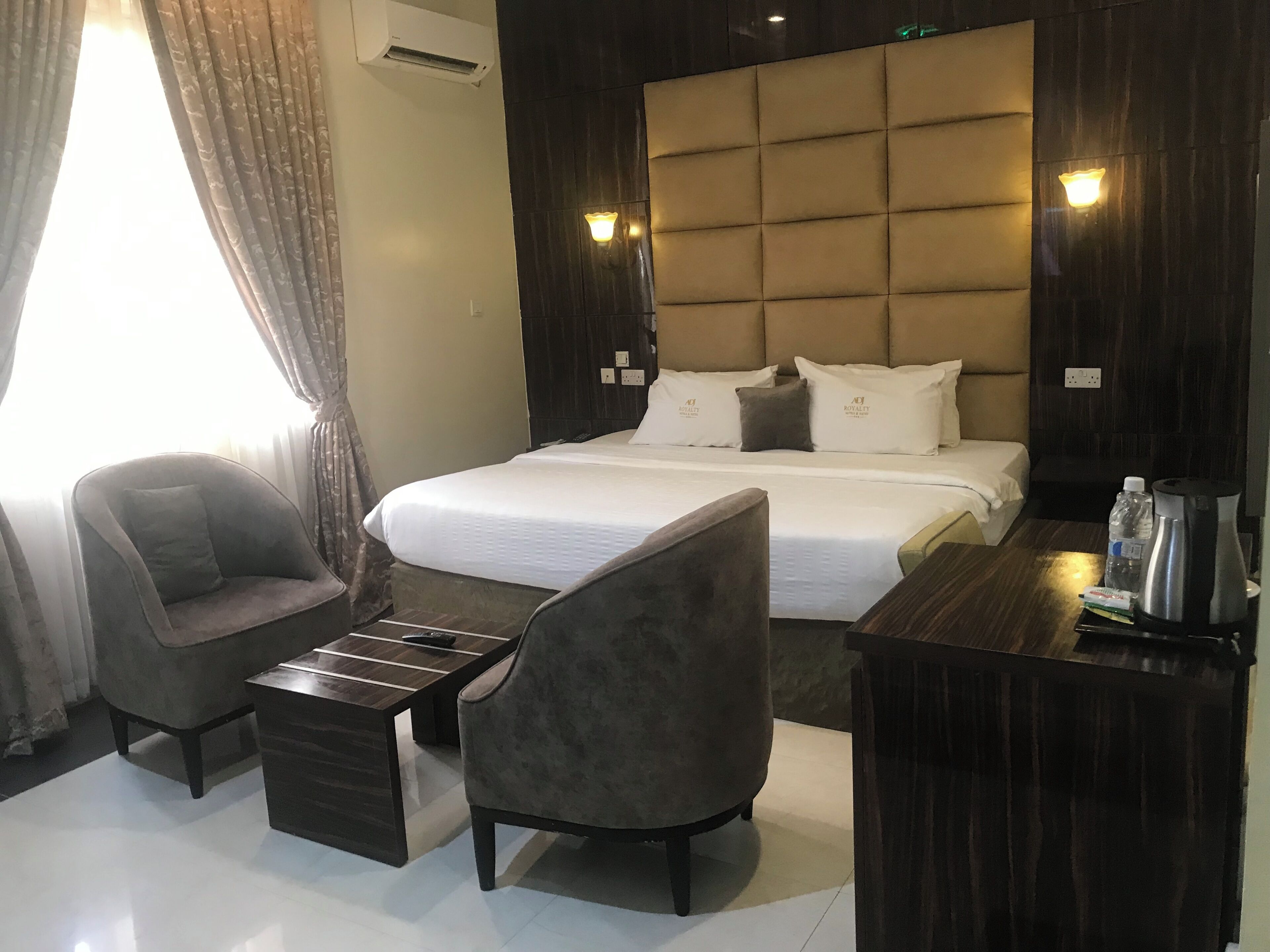 A hotel room with a large bed, two armchairs, a small table, and a bedside table with a kettle and water bottle.