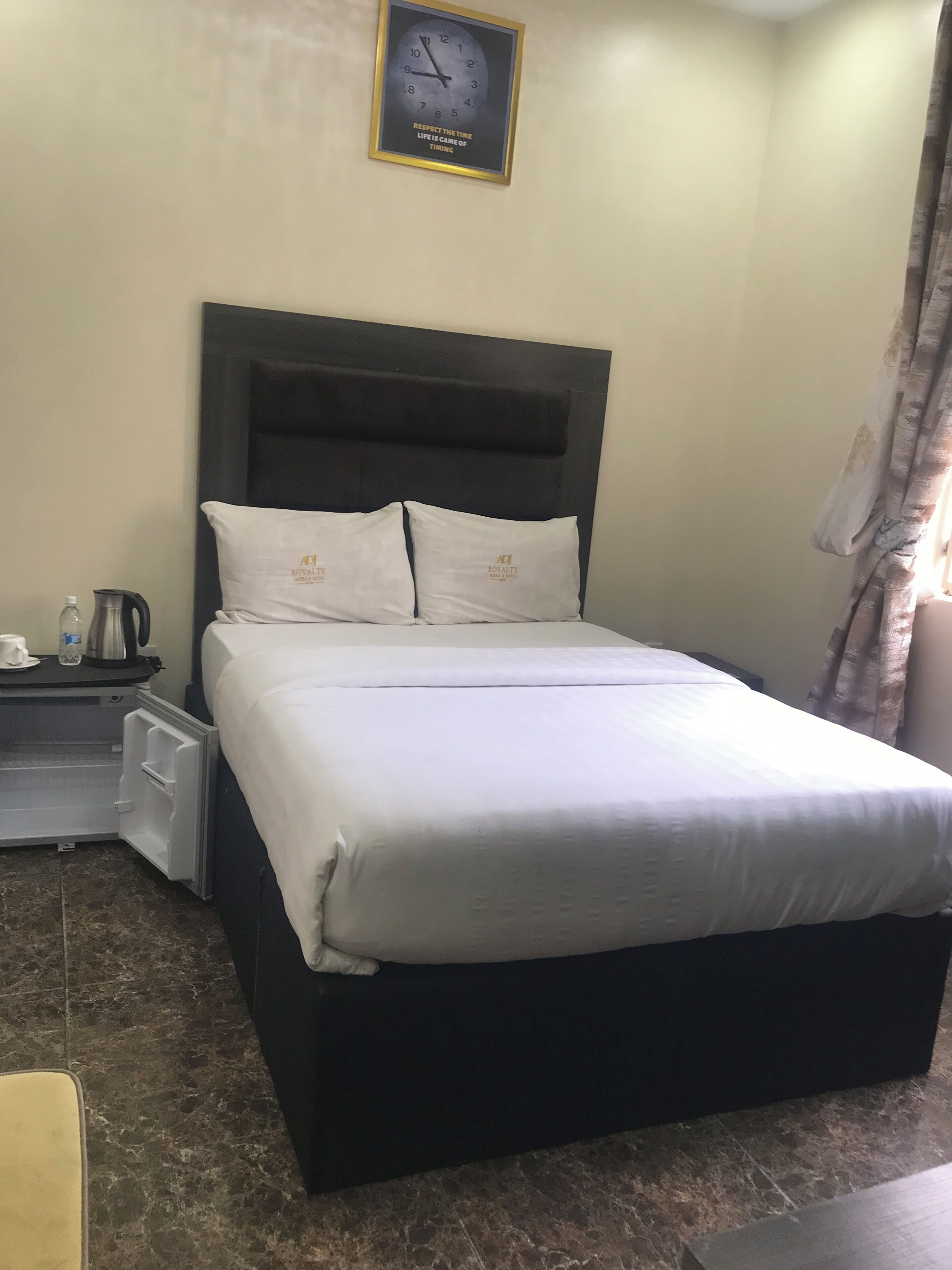 Una habitación de hotel con cama, mesita de noche, reloj y ventana con cortinas.