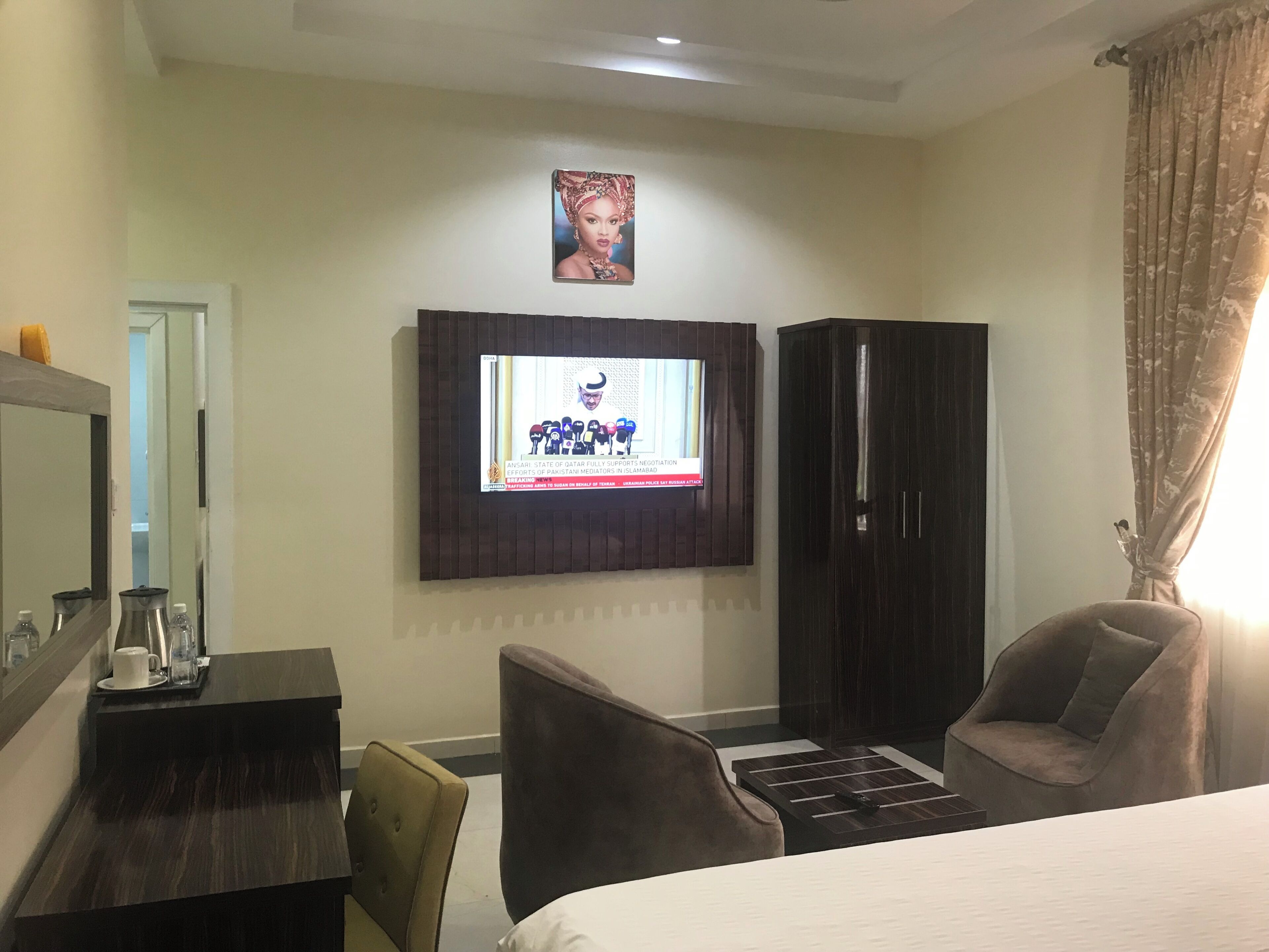 Habitación de hotel con televisión montada en la pared, cama, escritorio con una taza y una tetera, silla y armario.