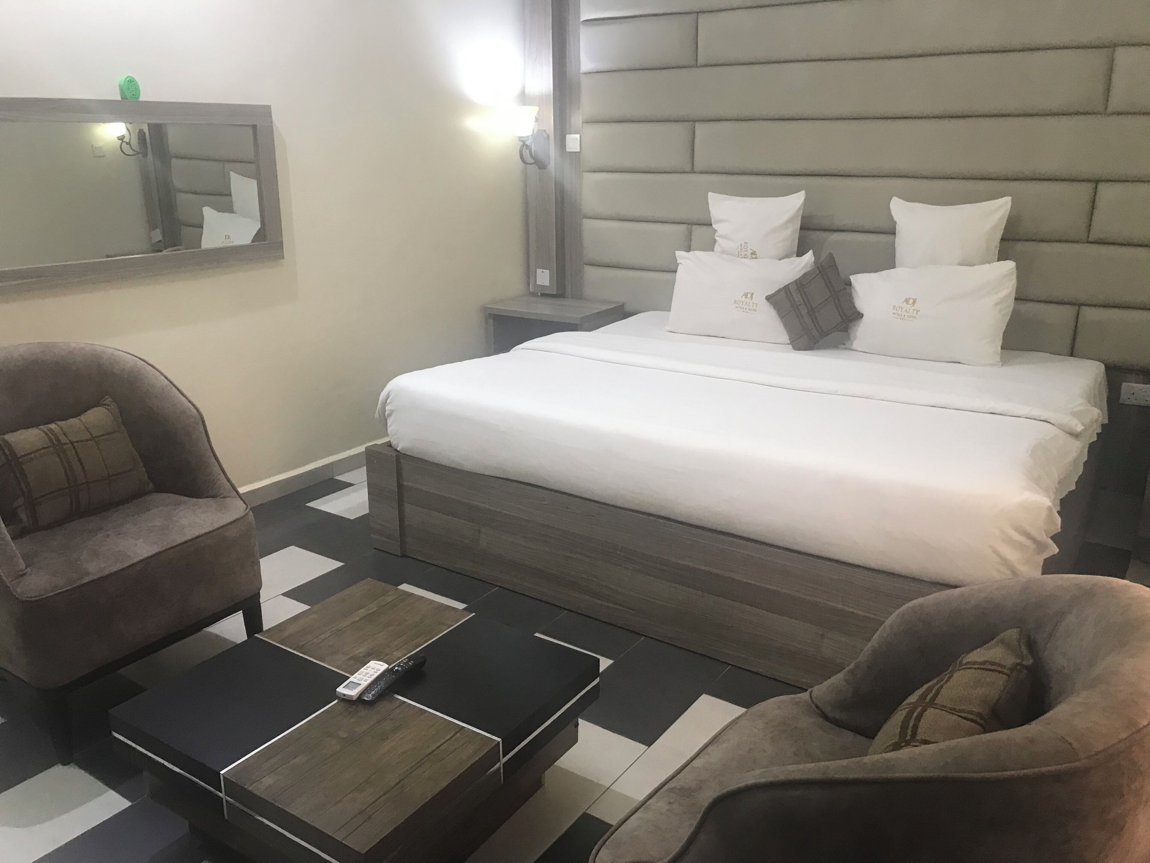 Quarto de hotel com uma cama, duas poltronas, uma mesinha de centro com um controle remoto, um criado-mudo e um espelho na parede.