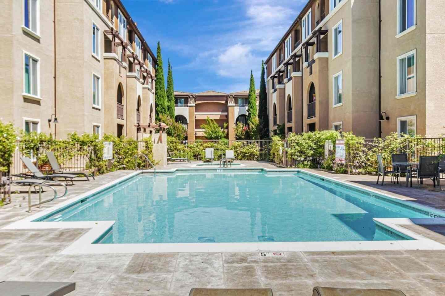 A rectangular swimming pool surrounded by lounge chairs and a building with multiple windows and balconies.
