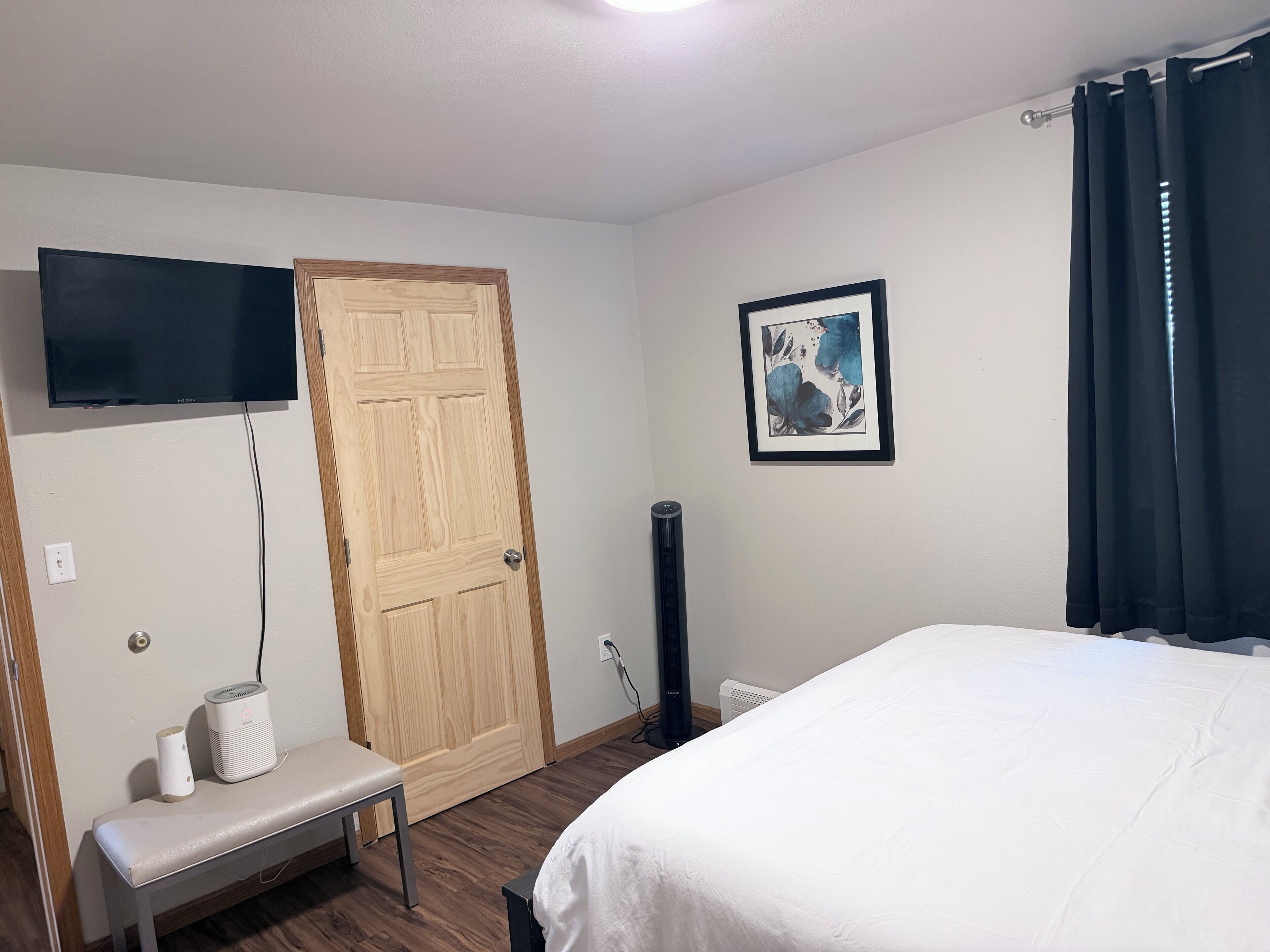 A hotel room with a bed, a bench, a television, a door, a radiator, and a picture on the wall.
