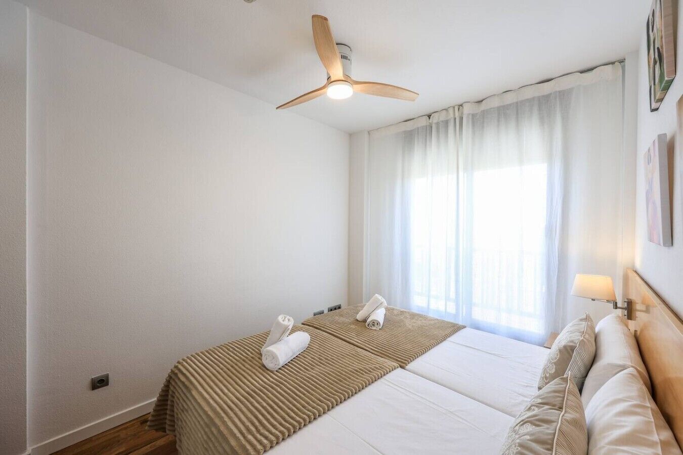 Una habitación de hotel con cama, ventilador de techo, ventana con cortinas y una lámpara de pared.