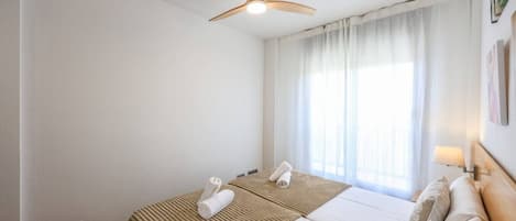 A hotel room with a bed, a ceiling fan, a window with curtains, and a wall-mounted lamp.