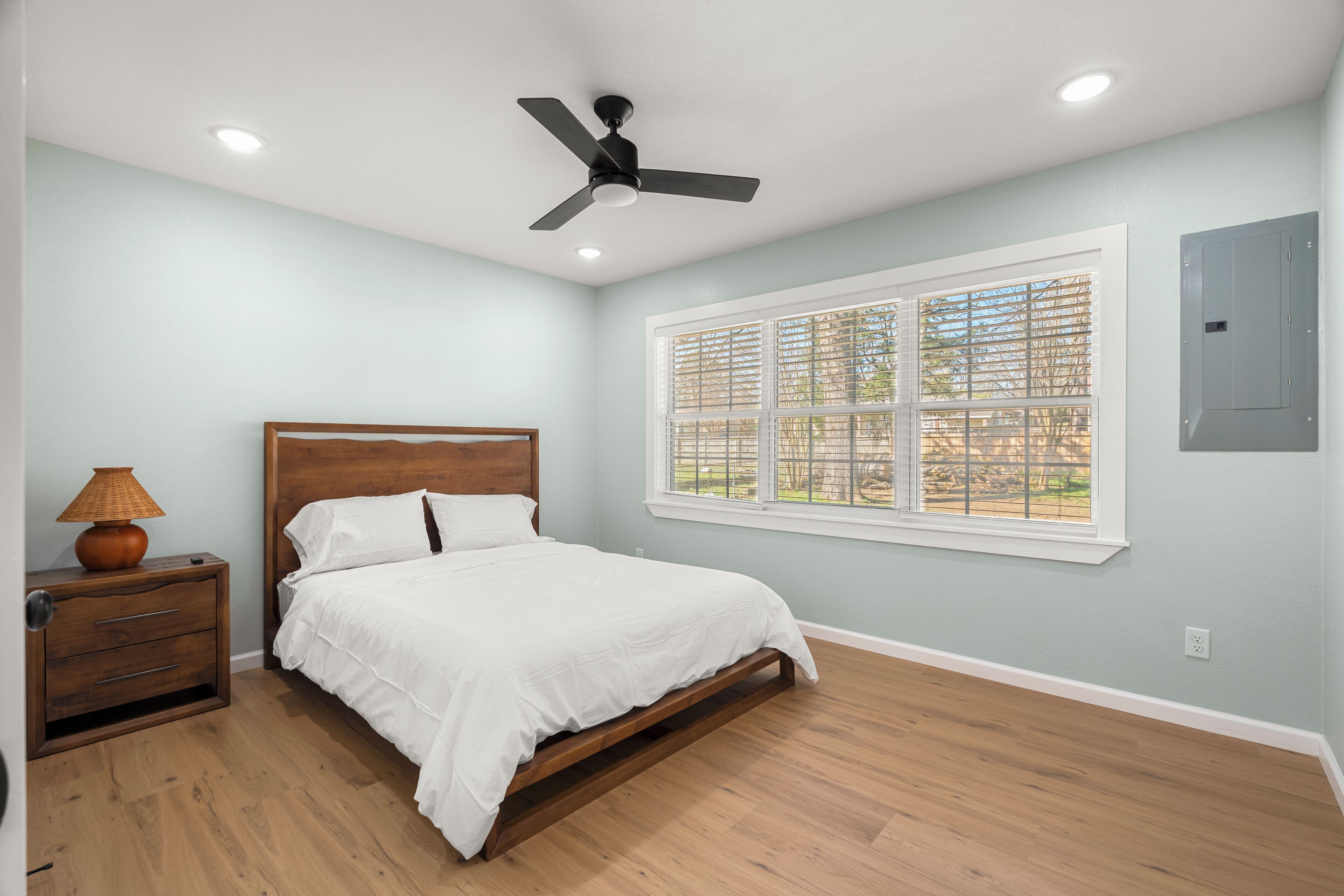 A bedroom with a bed, a nightstand, a lamp, a ceiling fan, and a window with blinds.