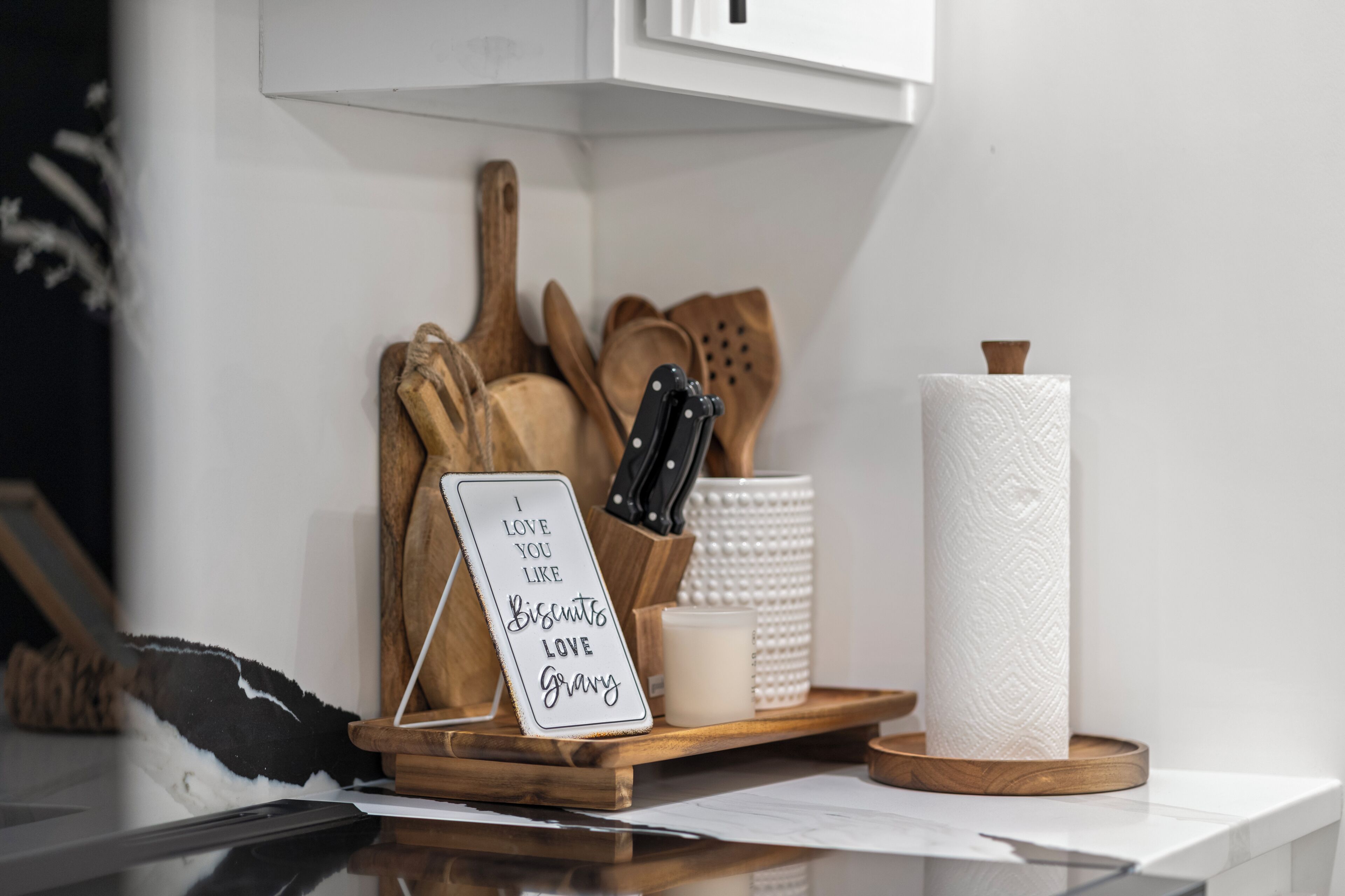 A kitchen counter with a sign that reads 'I LOVE YOU LIKE Biscuits LOVE gravy,' a roll of paper towels, a knife block, and wooden utensils.