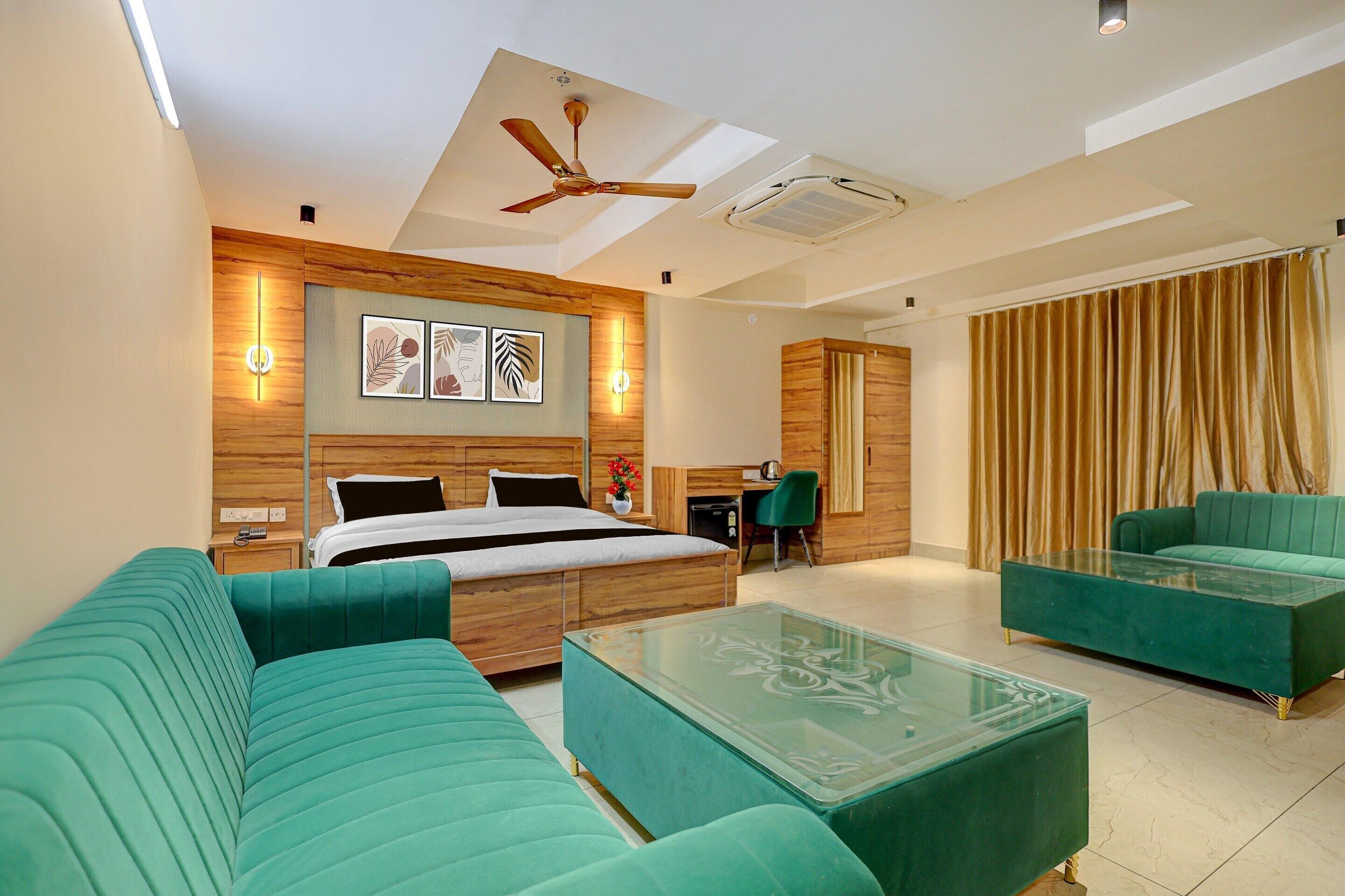 A modern hotel room with a bed, two green sofas, a glass coffee table, a desk with a chair, and a ceiling fan.