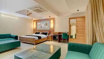 A modern hotel room with a bed, two green sofas, a glass coffee table, a desk with a chair, and a mirror.