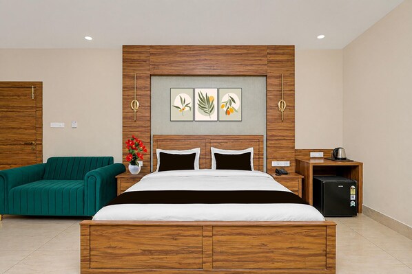 A hotel room with a wooden headboard, a bed with white bedding, a green sofa, a small table with a kettle, and a wall-mounted artwork.