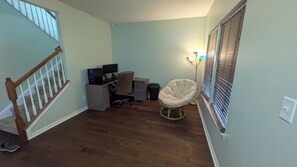 A room with a wooden staircase, a desk with two monitors, a chair, and a window with blinds.