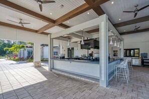 Ein Außenbereich mit Bar, Theke, Barhocker, Deckenventilatoren und Blick auf einen Garten.