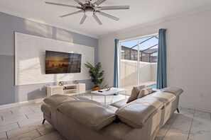 A modern living room with a sectional sofa, a flat-screen TV mounted on the wall, a ceiling fan, and a large window with curtains. - Family Friendly Home, Private Pool, Near Disney (Kissimmee)