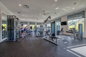 Ein großzügiger Fitnessraum mit verschiedenen Trainingsgeräten, großen Fenstern und einem Deckenventilator.