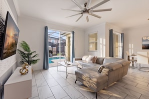 A modern living room with a sofa, a coffee table, and a television. There is a glass door leading to a pool area. - Family Friendly Home, Private Pool, Near Disney (Kissimmee)