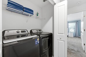 Une buanderie équipée de deux machines à laver et de deux sèche-linge à chargement frontal, d’une porte blanche et d’une fenêtre avec des rideaux.