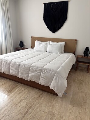 A bed with a white quilt, wooden headboard, and two bedside tables with lamps. - Modern apartment located in Aldea Zama with a pool, gym, and green spaces. (Tulum)