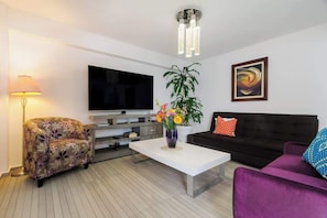 Ein modernes Wohnzimmer mit Flachbildfernseher, Couchtisch mit Blumenvase, Sofa und violettem Sessel.
