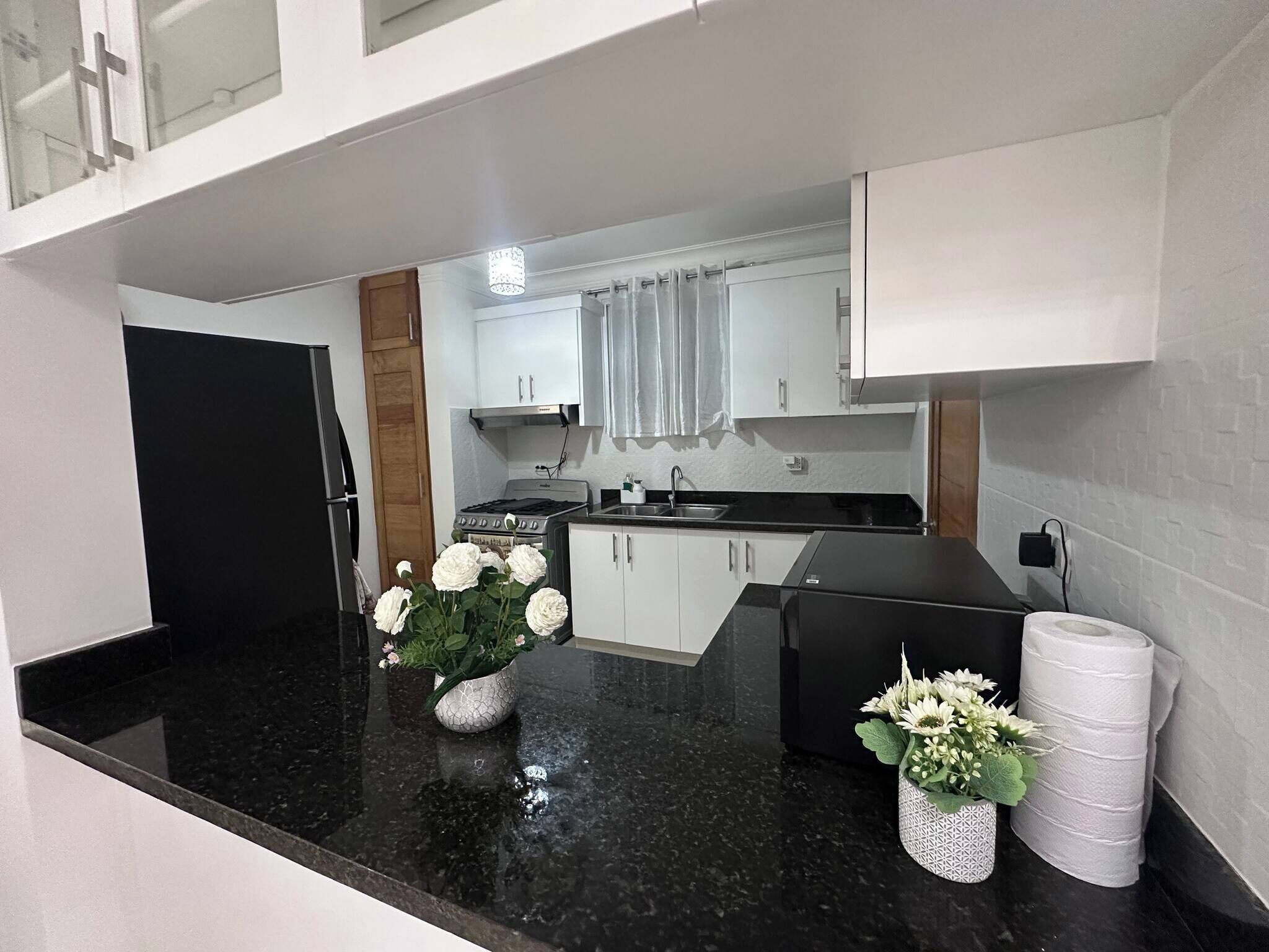 A compact kitchen with white cabinets, a black countertop, and a stove. There are two flower arrangements on the counter.