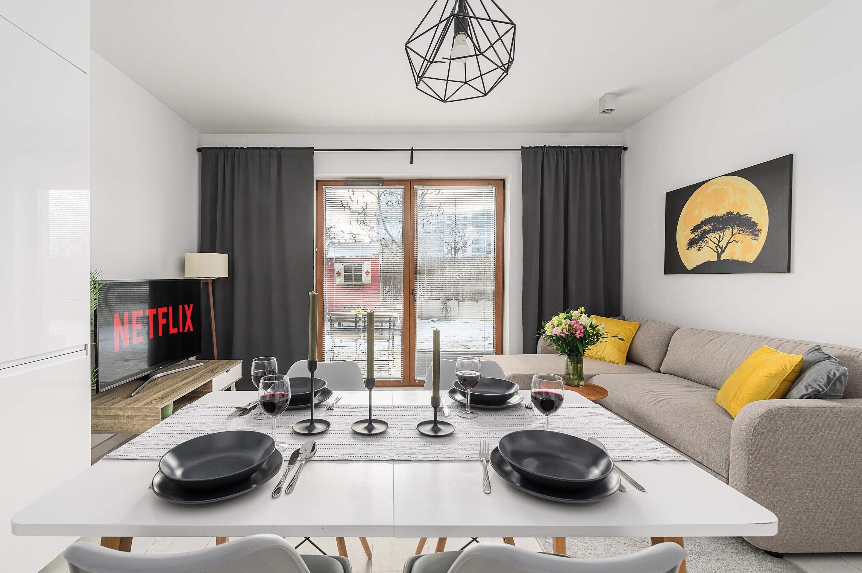 Un salon moderne comprenant un coin repas, un canapé avec des coussins jaunes, un tableau représentant un arbre et la lune, et un téléviseur affichant le logo de Netflix.